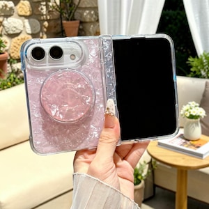 May include: A clear, foldable phone case featuring a light pink, pearlescent, crushed-shell texture. The case includes a matching circular, collapsible grip mount on the back and a cutout for a dual-camera system with a flash.
