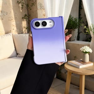 May include: A foldable smartphone case featuring a smooth gradient design that transitions from a deep purple at the top to a lighter lavender shade at the bottom. The case includes a rectangular cutout for the camera lenses and flash.
