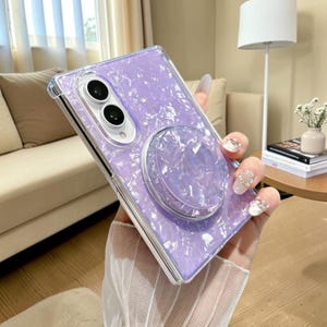 May include: A rectangular smartphone case featuring a glossy, iridescent light purple finish with a textured, crushed-shell pattern. The case includes a matching circular collapsible grip attached to the back and a cutout for a dual-lens camera system.