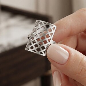 May include: A silver-toned metal ring featuring a rectangular face with a geometric lattice or grid cutout pattern. The ring has a smooth, polished finish and a thin band that connects to the openwork rectangular top section.