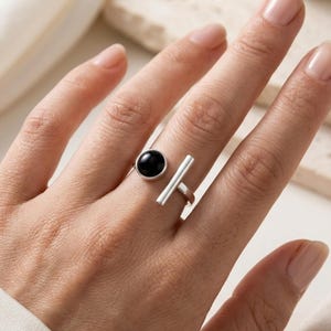 May include: A silver open-style ring featuring a smooth, round black stone on one end and a polished silver bar on the other. The ring is crafted from a metallic band that remains open at the top, showcasing a contrast between the dark circular gemstone and the sleek, straight silver element.