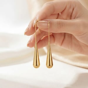 May include: A pair of gold-toned drop earrings featuring a smooth, elongated teardrop shape suspended from thin, curved wire hooks. The metal surface has a polished, reflective finish that catches the light, creating a bright, metallic appearance.
