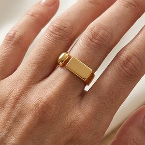 Könnte beinhalten: Ein goldfarbener Ring mit einem glatten, rechteckigen Stangenelement, das horizontal über dem Finger liegt, ergänzt durch einen kleinen, abgerundeten, polierten goldfarbenen Perlenakzent an der Seite des Rings für ein markantes Aussehen.