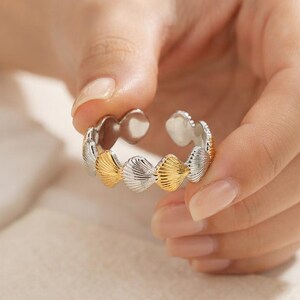Könnte beinhalten: Ein verstellbarer offener Ring mit einem sich wiederholenden Muster aus abwechselnd gold- und silberfarbenen Muschelmotiven. Das Band besteht aus einzelnen muschelförmigen Segmenten, die zu einem durchgehenden kreisförmigen Design verbunden sind.