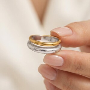 May include: A two-tone ring featuring a thin, polished gold-colored band stacked on top of a wider, smooth silver-colored band. The design consists of two distinct metal rings joined together to create a single layered piece of jewelry.