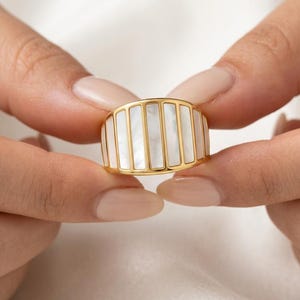 May include: A gold band ring featuring a series of vertical, rectangular white mother-of-pearl inlays set within a polished gold frame. The design consists of alternating gold bars and smooth, iridescent white stone segments arranged in a row.