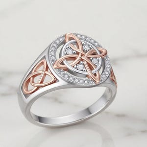 May include: A two-tone ring featuring a silver band and a circular centerpiece. The center displays a rose gold Celtic knot design surrounded by a halo of small clear stones. Additional rose gold Celtic knot motifs adorn the sides of the band.