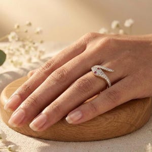 K&ouml;nnte beinhalten: Ein goldener Ring in Form einer gewundenen Schlange, besetzt mit kleinen klaren Steinen und zwei kleinen roten Steinen als Augen. Der Ring schlingt sich um den Finger, wobei der Schlangenkopf auf dem oberen Teil des Fingers aufliegt.