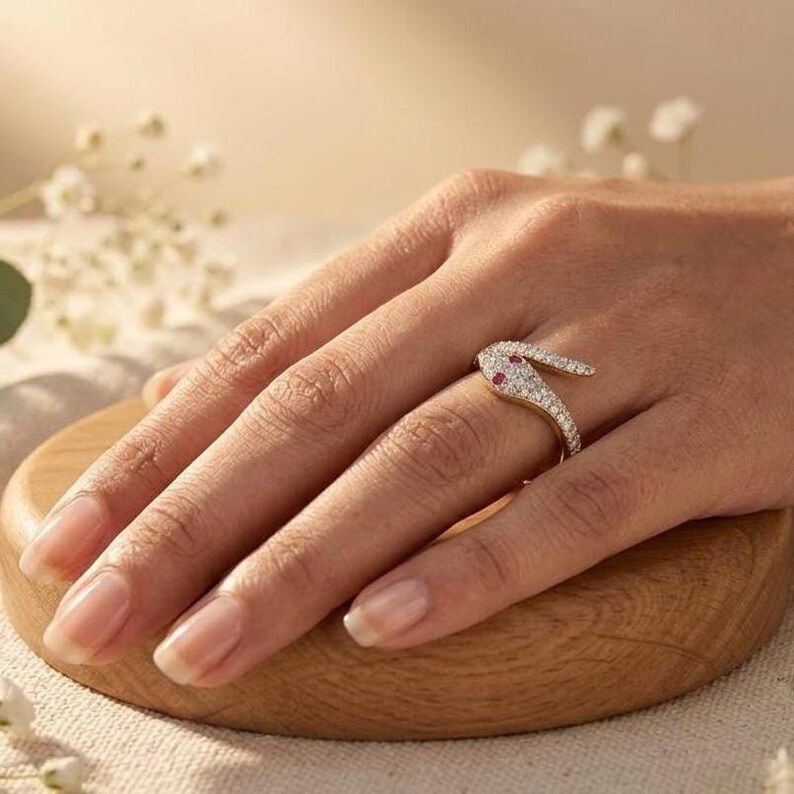 K&ouml;nnte beinhalten: Ein goldener Ring in Form einer gewundenen Schlange, besetzt mit kleinen klaren Steinen und zwei kleinen roten Steinen als Augen. Der Ring schlingt sich um den Finger, wobei der Schlangenkopf auf dem oberen Teil des Fingers aufliegt.