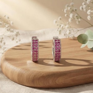 May include: A pair of silver hoop earrings featuring two rows of small, round, vibrant pink gemstones set along the outer surface. The earrings have a hinged closure mechanism and a polished metallic finish, showcasing a bright and reflective appearance.
