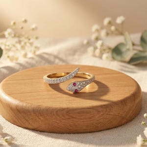 May include: A gold-toned snake-shaped ring featuring a pavé-set band with small clear stones and two small red stones serving as the eyes of the snake. The ring is designed in an open-ended style, allowing the head and tail to overlap slightly.