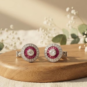 Puede incluir: Un par de gemelos redondos con una piedra central redonda transparente rodeada por un anillo de piedras rojas talla baguette, enmarcados por un borde de pequeñas piedras transparentes en un acabado de metal plateado.