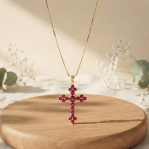 May include: A gold-toned necklace featuring a cross-shaped pendant adorned with multiple round, deep red gemstones. The pendant is suspended from a delicate, thin gold-toned chain, creating a refined and classic jewelry piece for any collection.