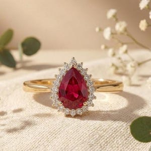 May include: A gold ring featuring a pear-shaped red gemstone center stone surrounded by a halo of small round white diamonds. The band is thin and polished gold, providing a classic look that highlights the vibrant red color of the central pear-cut stone.