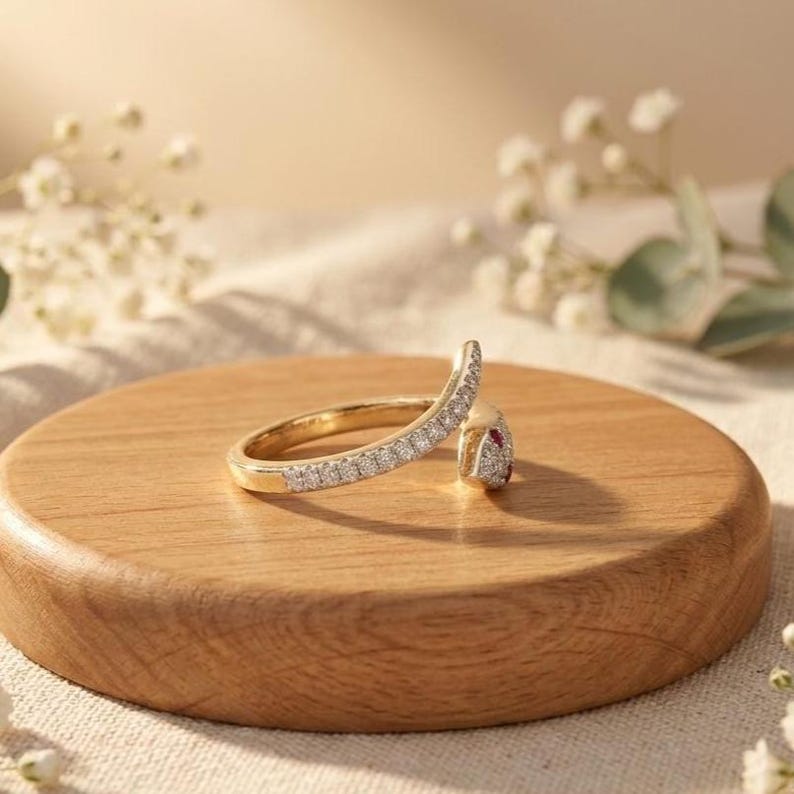 K&ouml;nnte beinhalten: Ein goldener Ring in Form einer gewundenen Schlange, dessen Ringschiene mit kleinen klaren Steinen besetzt ist und dessen Kopf einen einzelnen roten Stein aufweist. Der Ring ist auf einem runden Holzsockel platziert und zeigt seine feine Metallarbeit.