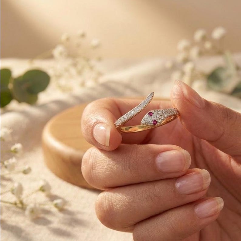 K&ouml;nnte beinhalten: Ein goldener Schlangenring mit einem pav&eacute;-gefassten Diamantk&ouml;rper und zwei kleinen roten Edelsteinaugen. Der Ring ist im Bypass-Stil gestaltet, wobei sich Kopf und Schwanz der Schlange &uuml;berlappen und so eine offene, verstellbare Ringschiene bilden.