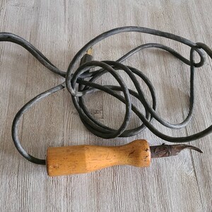May include: A vintage soldering iron with a wooden handle and a black electrical cord. The iron has a curved tip and a small metal loop on the handle.