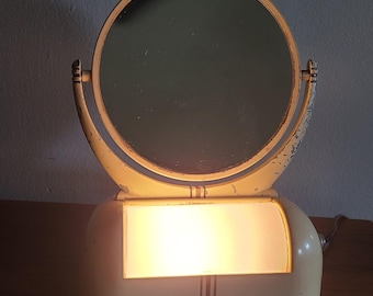 May include: A vintage vanity mirror with a light-up base. The mirror is round and has a cream-colored frame. The base is also cream-colored and has a rectangular light panel.