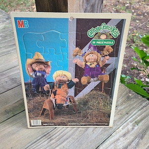 May include: A vintage Cabbage Patch Kids 25-piece puzzle featuring three Cabbage Patch Kids dolls. The puzzle is on a blue and brown background with a white border. The puzzle is for ages 3 to 5.