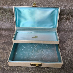May include: A vintage two-tiered jewelry box with a light blue interior and a cream-colored exterior. The box has a gold-colored latch and hinges.