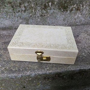 May include: A white jewelry box with a gold latch and a decorative floral design around the edge.