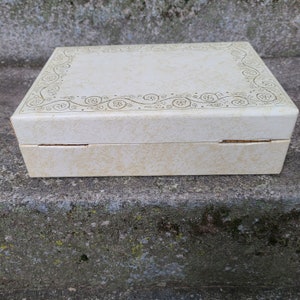 May include: A white rectangular box with a gold floral design around the edge of the lid. The box is closed and sitting on a gray concrete surface.