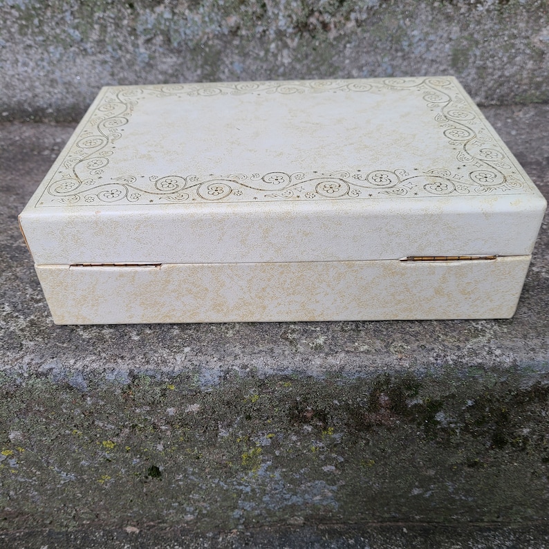May include: A white rectangular box with a gold floral design around the edge of the lid. The box is closed and sitting on a gray concrete surface.