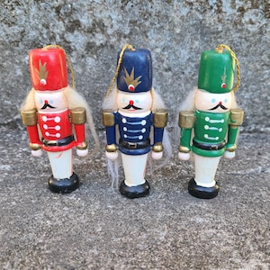 May include: Three wooden nutcracker ornaments, each with a different color scheme. The first is red with white accents, the second is blue with white accents, and the third is green with white accents. Each nutcracker has a white beard and a black mustache.