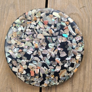 May include: A round, decorative coaster with a black base, inlaid with a mosaic of iridescent shell fragments in shades of white, pink, and green. The coaster is approximately 4 inches in diameter, suitable for holding a glass or mug.