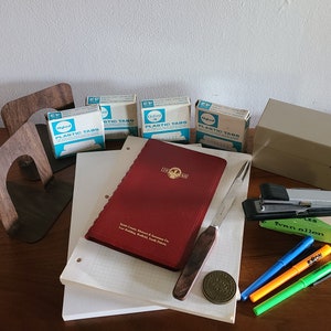 May include: A set of office supplies including a red notebook, a silver letter opener, a stapler, a box of plastic tabs, a wooden bookend, and a few pens and highlighters.