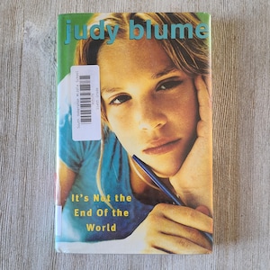 May include: A paperback book titled "It's Not the End of the World" by Judy Blume. The cover features a young person with a pen, and the title is in yellow and green. A library sticker is on the spine.