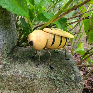 May include: A ceramic bee figurine with a smooth, pale yellow body featuring black horizontal stripes and a rounded head with black eyes. The insect has thin, metallic wire legs with black tips and a pair of pale yellow wings resting on its back.