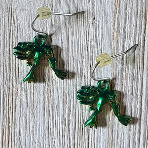 May include: A pair of green frog earrings with silver-tone ear wires. The frog charms have a metallic finish and are detailed with visible eyes and limbs. The earrings are displayed on a light-colored, textured wooden surface.