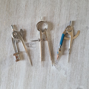 May include: Three silver drafting compasses with sharp points are arranged on a light wood-grain surface. One compass has a blue pencil holder. The tools are used for drawing circles and arcs.