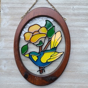 May include: A stained glass window hanging on a wooden frame. The design features a blue bird with yellow wings and a yellow flower with green leaves.