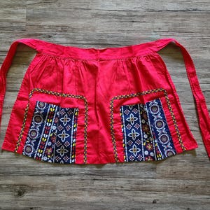 May include: A bright red apron with two pockets featuring a blue, white, and yellow patterned design. The apron has long red ties and a decorative trim around the pockets. The apron is likely for kitchen or craft use.