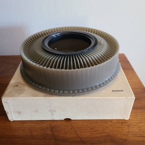 May include: A gray plastic Kodak slide carousel with a black ring around the center hole. The carousel has numbers 1 through 30 printed around the edge.