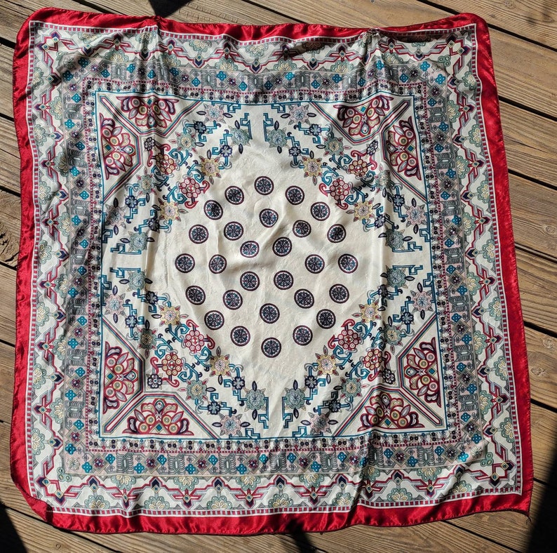 May include: A white silk scarf with a red border and a colorful geometric pattern. The pattern features a repeating design of circles, flowers, and geometric shapes.