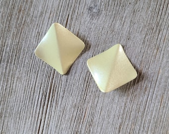 May include: Two pale yellow square earrings with a pyramid design. The earrings are set against a weathered, gray wooden surface. The earrings have a subtle sheen, reflecting light.