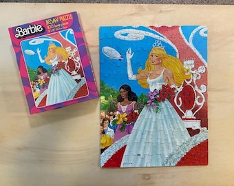 May include: A 100-piece jigsaw puzzle featuring a Barbie doll dressed in a white gown and a crown, standing on a red and white float with a blue sky background. The box for the puzzle is pink and white with the words "Barbie Jigsaw Puzzle" and "100 large pieces".