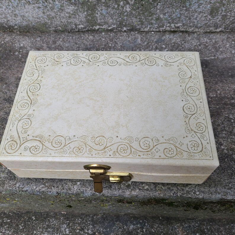 May include: A white jewelry box with a gold floral design and a brass latch.