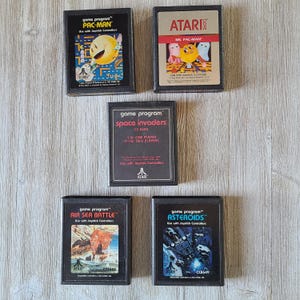 May include: Five vintage Atari game cartridges for the Atari 2600 console. The cartridges are black with white text and graphics. The games are Pac-Man, Ms. Pac-Man, Space Invaders, Air Sea Battle, and Asteroids.