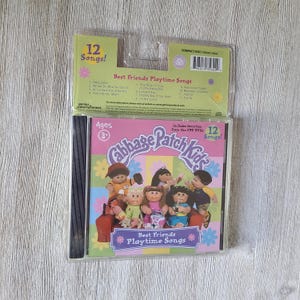 May include: A Cabbage Patch Kids "Best Friends Playtime Songs" CD in a clear plastic case. The CD cover features illustrations of Cabbage Patch Kids dolls. The packaging includes the text "12 Songs!" and "Ages 3+".