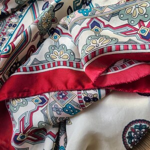 May include: A white and red patterned scarf with a geometric design. The scarf has a red border and a white background with intricate patterns in red, blue, and green.