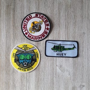May include: Three embroidered patches: a circular patch with "UNITED STATES MARINES" text and a bulldog graphic, a circular patch with a panther graphic and "ANVIS HUD" text, and a rectangular patch with a green helicopter and "HUEY" text.