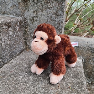 May include: A plush brown monkey toy with a light brown face, ears, hands, and feet. The stuffed animal has a soft, furry texture and is standing on a stone surface. The toy has the brand name "Aurora" on a tag.