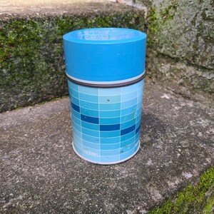 May include: A cylindrical Thermos food jar with a bright blue plastic lid. The body features a grid pattern of various shades of blue, ranging from light sky blue to deep navy. The brand name Thermos is printed in white near the bottom edge.