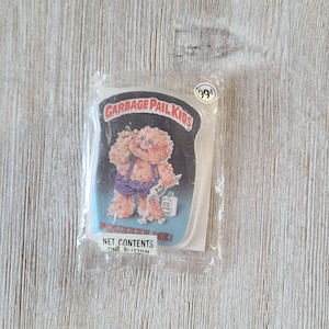 May include: A Garbage Pail Kids button featuring a character with a blue shirt and a bandage on their nose. The button is packaged in a clear plastic bag with a price tag of 99 cents. The text on the button reads "Garbage Pail Kids" and "Squeeze Me!"  The text on the packaging reads "Net Contents One Button."
