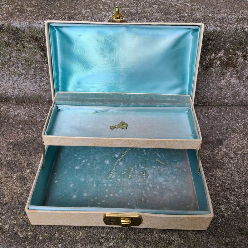 May include: A vintage two-tiered jewelry box with a light blue interior and a cream-colored exterior. The box has a gold-colored latch and hinges.