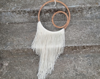 May include: A white macrame wall hanging with two wooden hoops. The hoops are concentric circles, with the smaller hoop inside the larger hoop. The macrame is made of long, thin strands of yarn that hang down from the hoops.